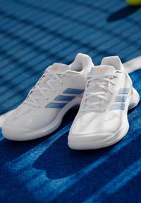 Λευκά αθλητικά παπούτσια Adidas με μπλε ρίγες τοποθετημένα σε μπλε υφή γηπέδου αθλημάτων κάτω από το φως του ήλιου.