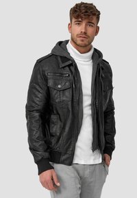 Black leather bomber jacket with a gray hood, featuring two chest pockets, ribbed cuffs, and a zip front. Worn over a white turtleneck.