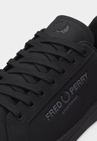Baskets Fred Perry noires avec lacets, montrant le logo de la marque et le texte "Sportswear" sur le côté ainsi que l'emblème de la couronne de laurier sur la languette.