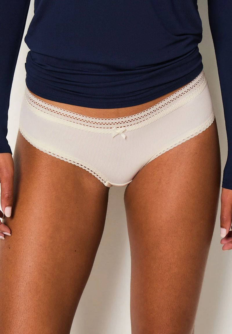 Slip hipster beige con texture a coste, bordino in pizzo in vita e un piccolo fiocchetto nella parte frontale. Tessuto morbido, design a vita media.