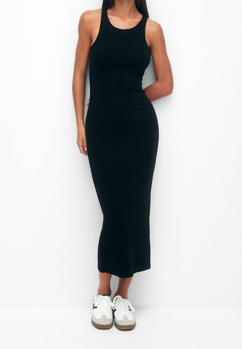 Robe midi noire sans manches en tissu lisse et extensible avec une silhouette ajustée. Associée à des baskets blanches et grises.