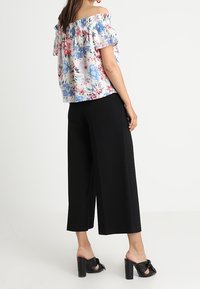 Donna che indossa un top floreale off-the-shoulder, pantaloni cropped neri a gamba larga e sandali con tacco alto neri, in piedi davanti a uno sfondo neutro.