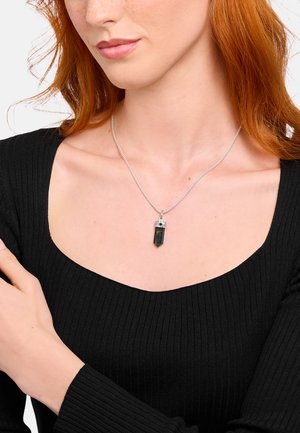 Frau mit roten Haaren, die ein schwarzes Top und eine silberne Kette mit einem dunklen Kristallanhänger trägt.