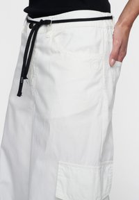 Witte cargo pants met een zwarte trekkoord in de taille, met een grote zijzak en geborduurd nummer "10" op de rechter dij.
