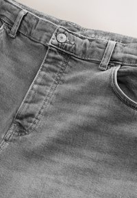 Primo piano di jeans denim grigi che mostrano la tasca anteriore, i passanti per cintura e il bottone metallico con la scritta "Premium Denim Goods".