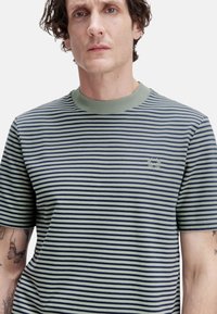 Camiseta a rayas en verde oliva y azul marino, con cuello redondo acanalado y un pequeño logo bordado en el lado izquierdo del pecho.