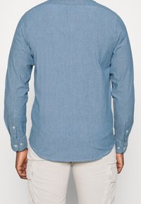 Camicia di denim azzurro chiaro con maniche lunghe, polsini con bottoni, orlo arrotondato e texture sottile. Vista posteriore senza dettagli aggiuntivi.