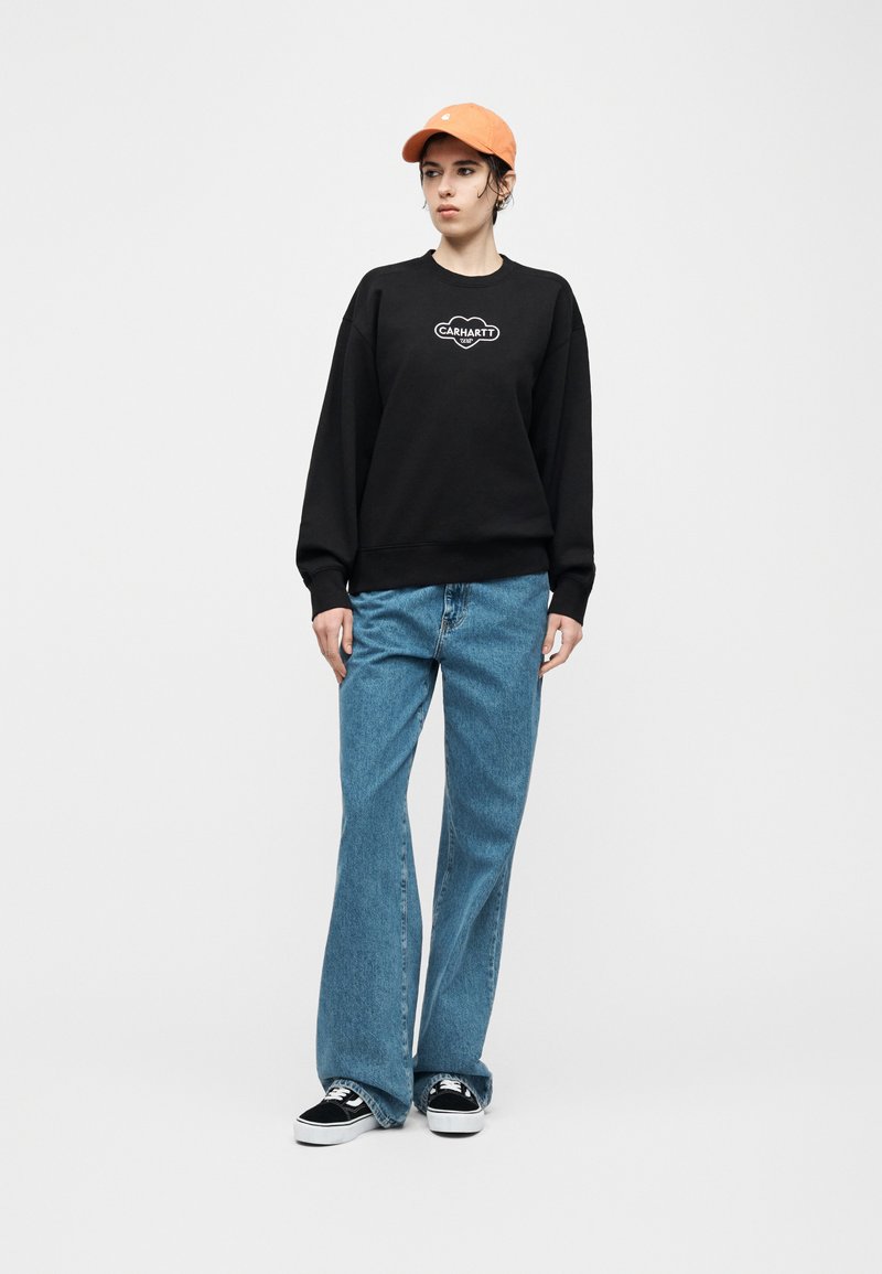 Giovane che indossa una felpa nera Carhartt, jeans a gamba larga azzurri, sneakers nere e bianche e un berretto arancione su sfondo grigio uniforme.