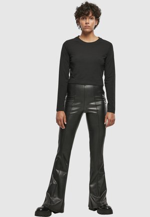 Nainen, jolla on lyhyet kiharat hiukset, musta pitkähihainen paita, mustat korkea vyötäröiset tekonahkaiset liehuvat housut ja mustat massiiviset saappaat.