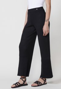 Pantalons larges à côtes noires avec une taille haute et un ourlet fendu, assortis de sandales noires à clous. La texture est légèrement extensible.