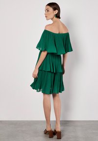 Grünes, plissiertes Off-Shoulder-Kleid mit geschichteten Rüschen, knielang und mit glatter Textur. Braune Blockabsatzsandalen runden das Outfit ab.