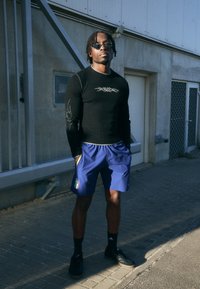 Camisa preta de mangas longas com desenhos prateados, calções azuis com a bandeira da Itália, meias pretas e ténis pretos. Cenário ao ar livre com um fundo de betão.