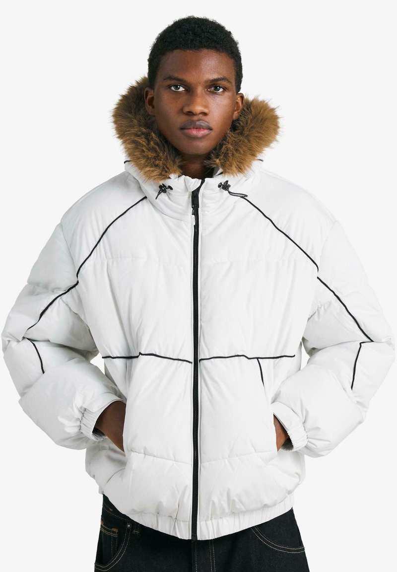 Biela prešívaná bunda s čiernym prešívaním, kapucňou lemovanou kožušinou a elastickými manžetami. Má zipsové zapínanie a boxovitý strih.