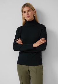 Zwarte coltrui van zacht materiaal, met ribgebreide manchetten en zoom, gecombineerd met olijfgroene op maat gemaakte broek met gouden accenten.