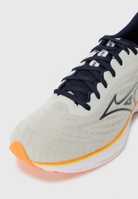 Chaussure de course avec une tige en tricot gris clair, des lacets navy et un accent orange sur la semelle. Présente une surface texturée et un design épuré.