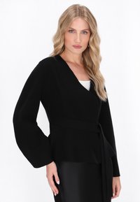 Jachetă neagră, înfășurată, dintr-un material moale, cu mâneci lungi în formă de clopot, un cordon la talie și un decolteu în formă de V. Textură netedă pe toată suprafața.
