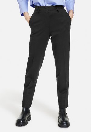 Persoon draagt op maat gemaakte zwarte broek, blauw overhemd en zwarte enkellaarzen met dikke zool, staat met handen in de zakken.