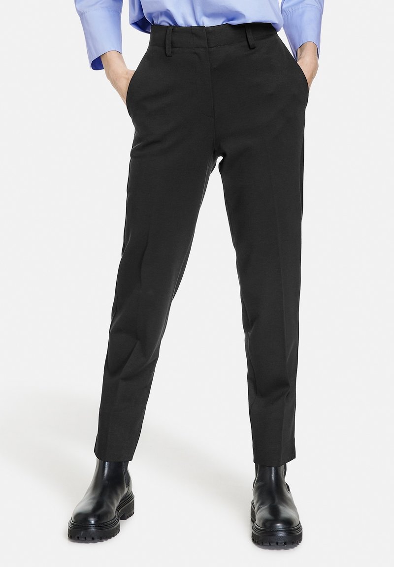 Personne portant un pantalon noir sur mesure, une chemise bleue et des bottines noires à semelle épaisse, debout les mains dans les poches.