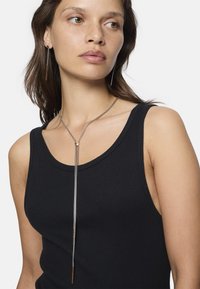 Zilveren ketting met een dunne, langwerpige bar pendant, gedragen over een zwarte geribbelde tanktop. De pendant heeft een kleine ronde accent.