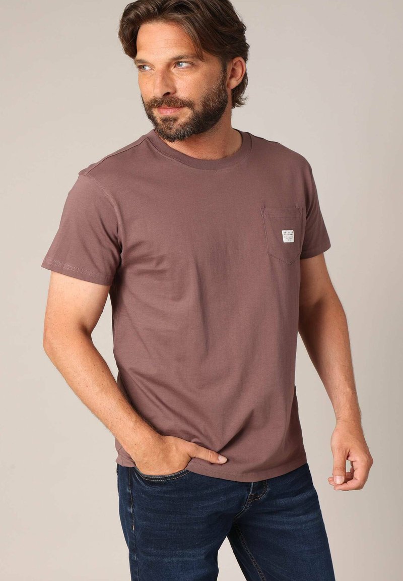 Man met een baard, gekleed in een bruine T-shirt met korte mouwen en een borstzak, en donkerblauwe spijkerbroek, kijkt naar links met één hand in zijn zak.