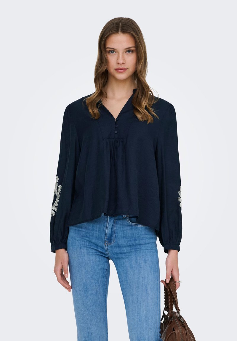 Jeune femme aux longs cheveux ondulés portant un chemisier ample bleu marine avec des broderies blanches sur les manches, un jean bleu clair, et tenant un sac à main marron.