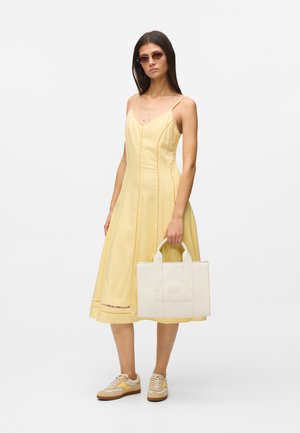 Frau trägt ein gelbes ärmelloses Midikleid, beige Sneaker, Sonnenbrille und hält eine weiße rechteckige Tragetasche mit eingeprägtem Text.