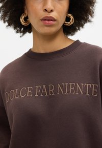 Sweatshirt castanho com decote canelado, apresentando "DOLCE FAR NIENTE" bordado em bege na parte da frente. Tecido de textura suave.