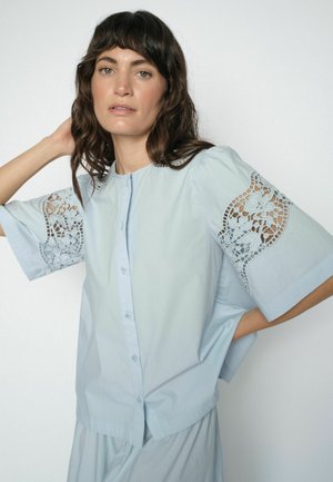 Žena s vlnitými tmavými vlasmi nosí svetlomodrú blúzku s gombíkmi a kvetinovými čipkovými výrezmi na rukávoch, pózuje s jednou rukou za hlavou.