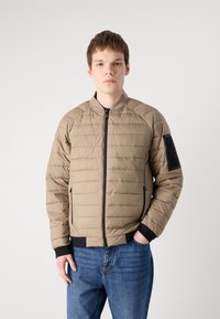 Casaco puffer bege com costuras horizontais, fecho preto, punhos canelados e um bolso com fecho no braço esquerdo. Usado sobre jeans azuis.
