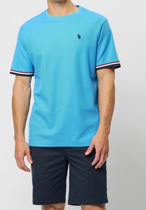Man draagt een felblauw T-shirt met korte mouwen met gestreepte mouwranden en marineblauwe korte broek, staat met ontspannen armen.