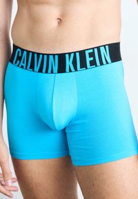 Svjetloplave pamučne bokserice imaju crni pojas s natpisom "CALVIN KLEIN" u podebljanoj cijan boji. Glatka tekstura, uski kroj.
