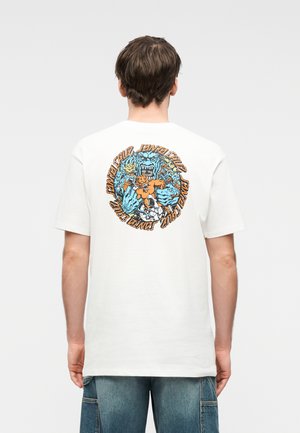 Mand klædt i hvid t-shirt med blå og orange grafik af et brølende monster, der holder en mindre figur, omgivet af stiliseret tekst.