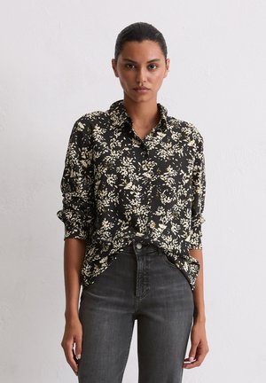 Camicia nera a bottoni con motivo floreale bianco; vestibilità rilassata, maniche lunghe arrotolate, abbinata a jeans grigio scuro. Tessuto dalla consistenza morbida.