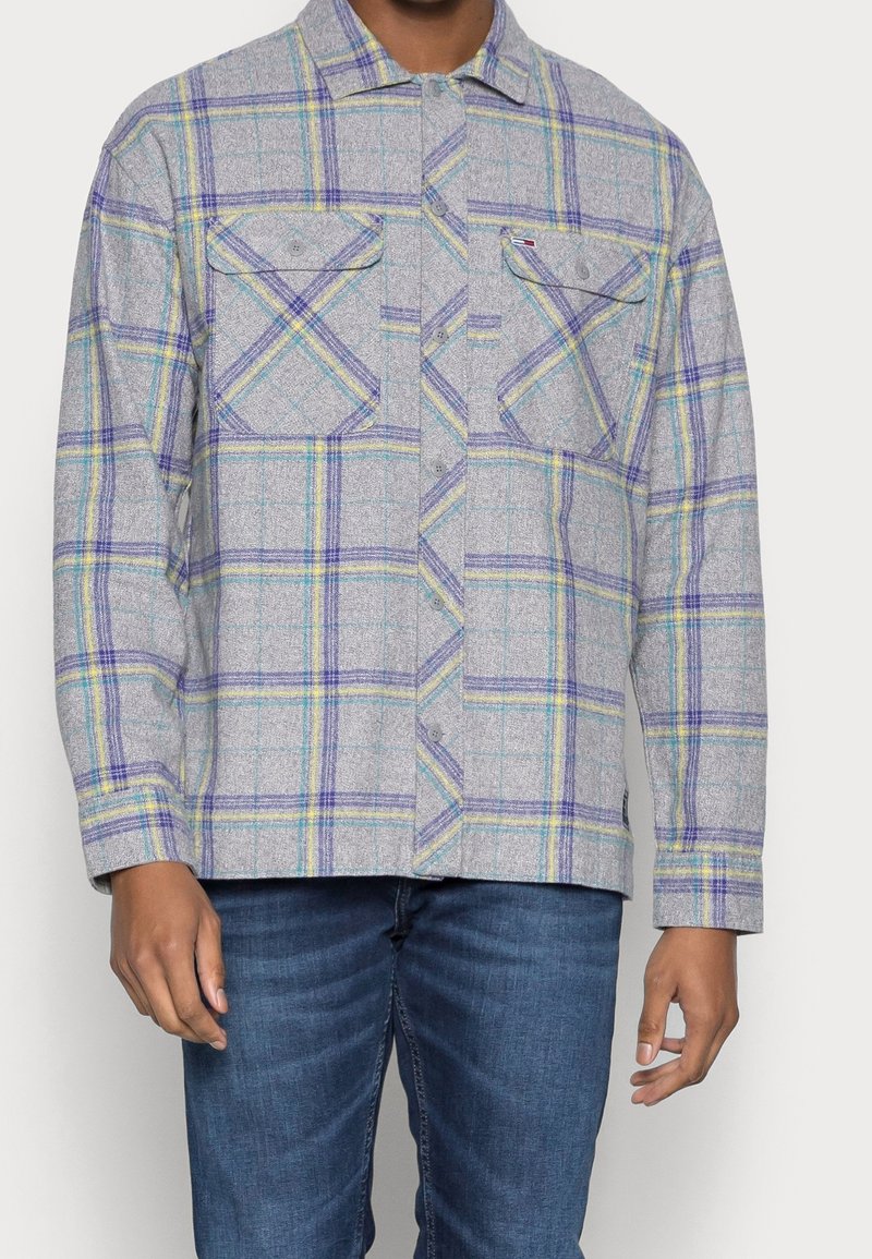 Uomo che indossa una camicia a quadri grigia chiara con righe viola, gialle e blu, abbinata a jeans blu, in piedi davanti a uno sfondo bianco.
