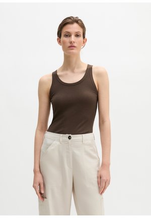 Kobieta o krótkich brązowych włosach, ubrana w brązowy, bezrękawnik z prążkowanego materiału i beżowe spodnie z wysokim stanem, stojąca na tle jednolitej białej ściany.