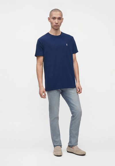 Άνδρας που φοράει μπλε σκούρο κοντομάνικο t-shirt, ανοιχτό μπλε τζιν και μπεζ μοκασίνια, στέκεται μπροστά σε ένα μονόχρωμο λευκό φόντο.