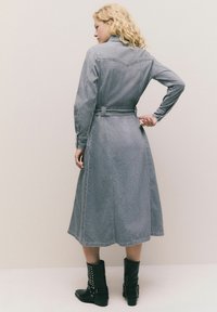 Frau mit lockigem blonden Haar, die ein graues Denim-Midikleid mit Gürtel und schwarze, mit Nieten besetzte ankle boots trägt, sie steht von der Kamera abgewandt vor einem schlichten Hintergrund.