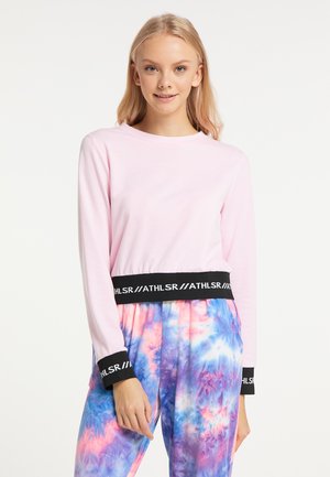 Blondine vrouw met een lichtroze longsleeve top met zwarte ATHLSR manchetten en tailleband, gecombineerd met kleurrijke tie-dye broeken.