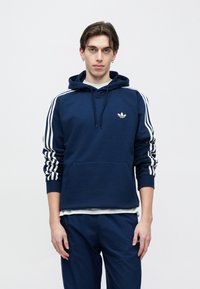 Marineblauwe capuchontrui met wit driestripes-design op de mouwen, kangoeroezak en klein wit logo op de borst. Zacht, glad oppervlak.