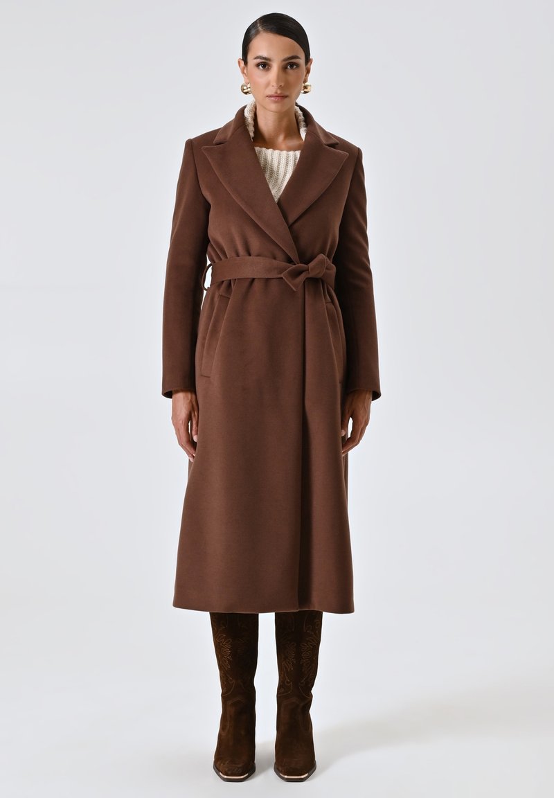Cappotto in misto lana marrone con vita regolabile, maniche lunghe e rever a punta. Maglione crema strutturato sotto e stivali marroni coordinati.