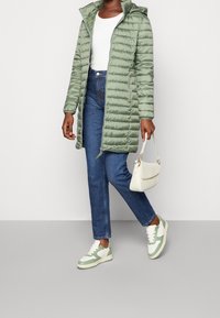 Grøn puffjakke med vandret syninger, hvid ribbet top, blå lige jeans og hvid sneakers med grønne nuancer. Lille hvid håndtaske.