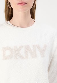 Balts vilnas krekliņš ar teksturētu, gaiši bežīgu "DKNY" logotipu. Dizainam ir apaļš kakla izgriezums un mīksta, pūkaina tekstūra.