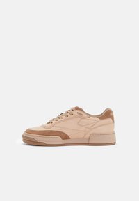 Beige athletic sneakers with suede and leather materials, featuring a textured sole, lace-up design, and subtle branding on the heel.