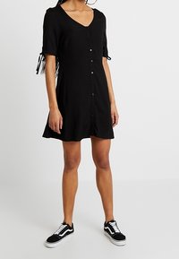 Abito nero a camicia con maniche corte, scollo rotondo e dettagli con laccio sulle maniche. Indossato con sneakers nere. Design semplice e casual.