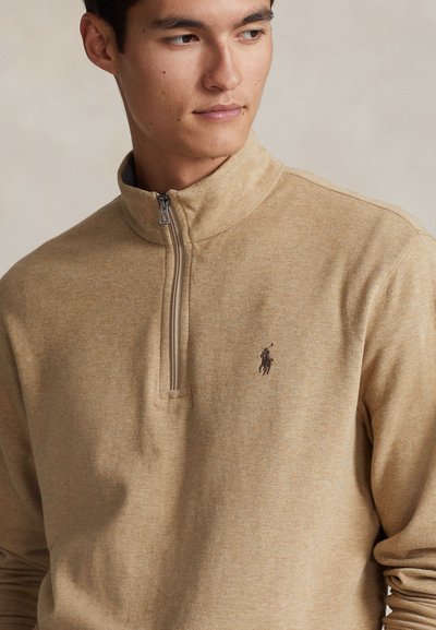 Mlad moški nosi bež pulover s četrtinskim zadrgo in majhnim temnim logotipom konja in jahača na levi strani prsnega koša, gleda vstran.