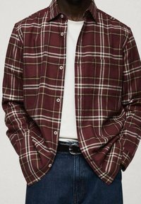 Camisa de cuadros en burdeos y blanco, con un diseño de botones, mangas largas y un corte relajado. Llevada sobre una camiseta blanca y unos jeans de mezclilla.