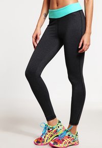 Leggings sportivi neri con una vita turchese, caratterizzati da una texture liscia e un design aderente. Abbinati a sneakers a fantasia colorata.
