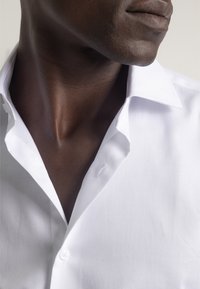 Camisa blanca de vestir con tejido texturizado, que presenta un collar clásico y un solo botón en el cuello. Acercamiento a la zona del collar.