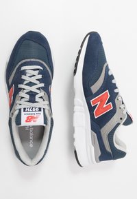 Marinblå sneakers med grå detaljer och röd New Balance-logotyp. Tillverkade av mesh och mocka, med en vit och svart gummisul.