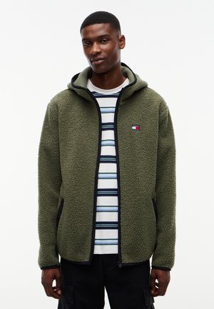 Young man wearing a green zip-up fleece jacket over a white and blue striped shirt standing against a plain white background.